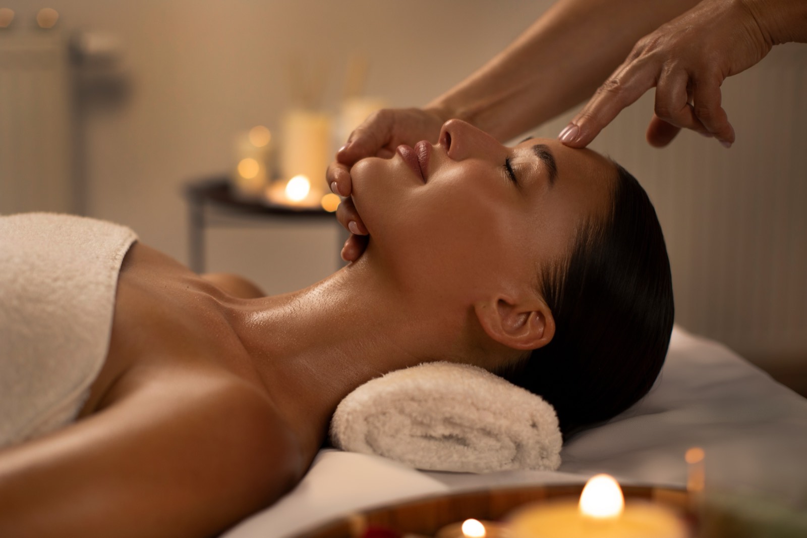 A client lying on a massage table with her eyes closed while a therapist works gently at her temples, in warm candlelight.